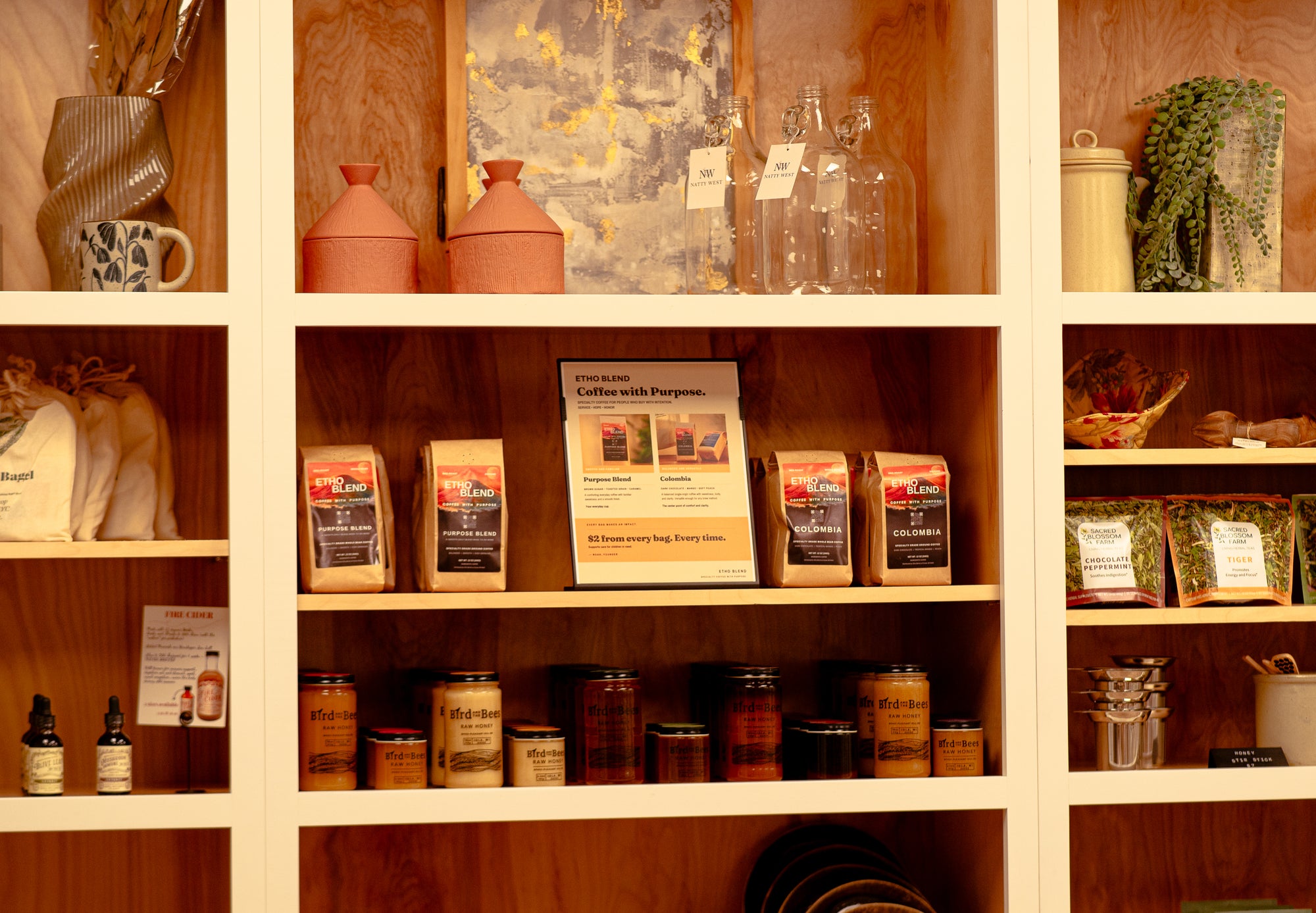 Wooden display shelves in a shop setting, holding packaged coffee bags, glass bottles, jars, ceramic containers, and small decorative items.