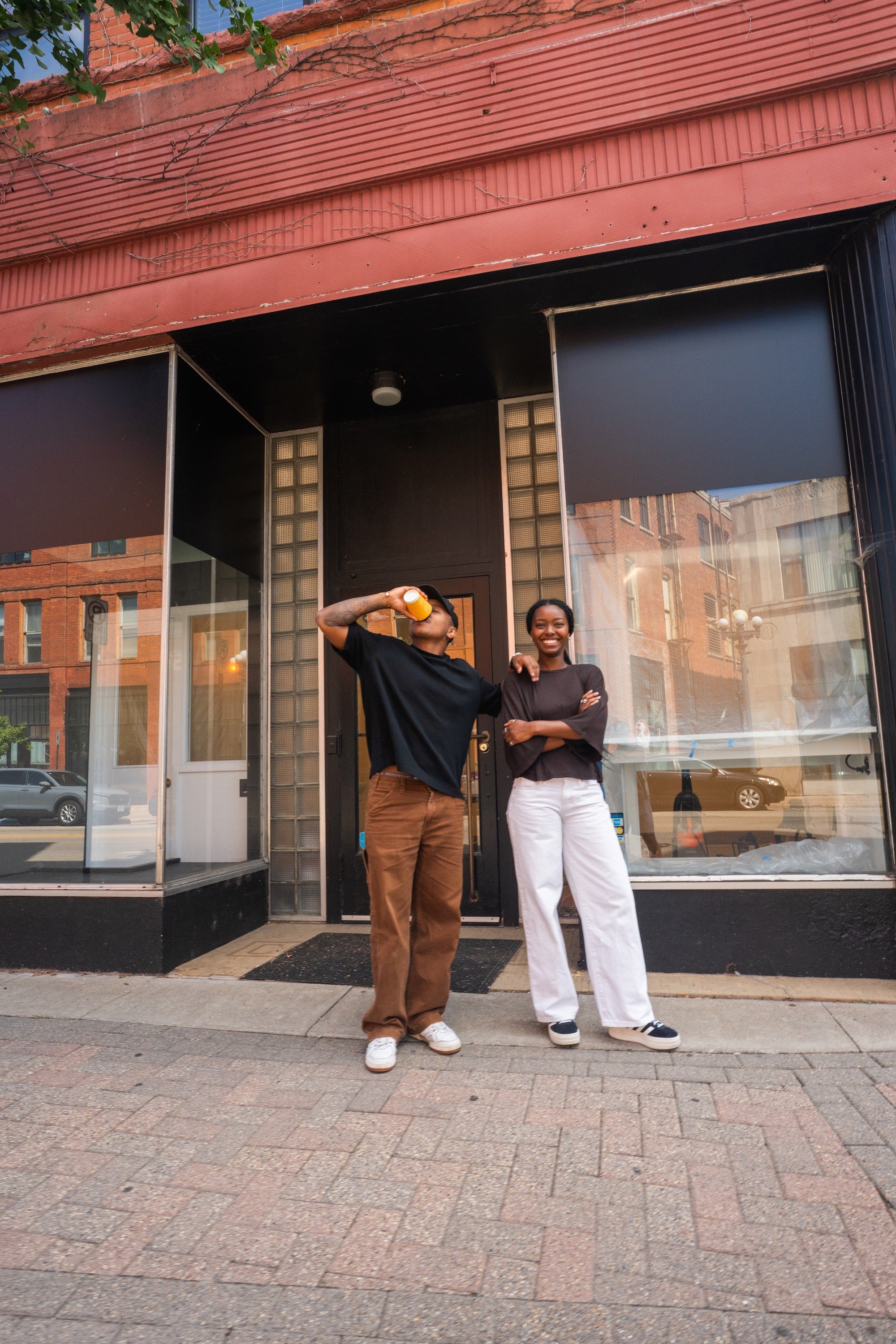Two people standing outside a storefront with large windows and a red brick building facade. The person on the left wears a black shirt, brown pants, and white shoes while holding a yellow cup to their mouth.